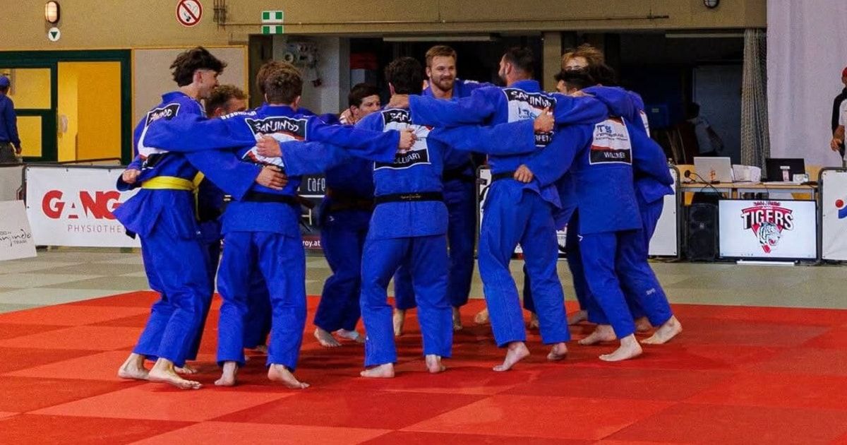 Eine Gruppe von Männern in blauen Kampfsportuniformen steht zusammen auf einem roten Teppich, wahrscheinlich in einem Fitnessstudio. Sie scheinen sich in einer Runde zu sammeln, möglicherweise in Vorbereitung auf einen Wettkampf oder Training.