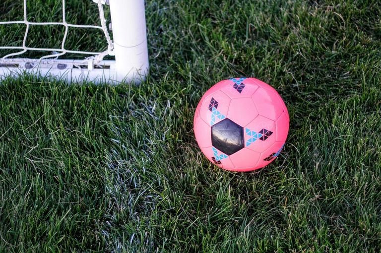 Ein rosafarbener Fußball mit geometrischen Mustern liegt auf dem grünen Rasen, in der Nähe eines Fußballtors.