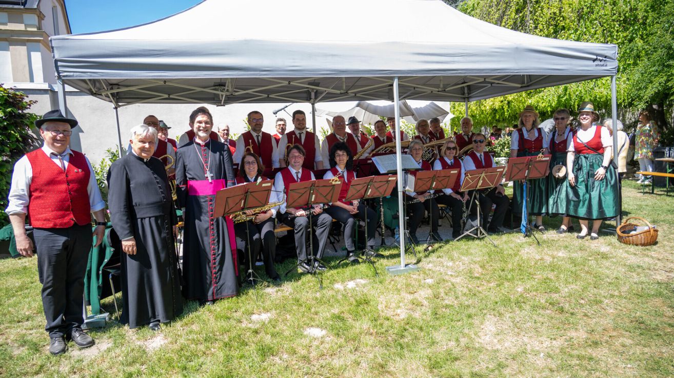 Eine Gruppe von Musikern in roten Westen sitzt unter einem weißen Zelt mit Notenständern vor ihnen. Eine Person in einem schwarzen Gewand und rotem Schärpe steht vor ihnen.