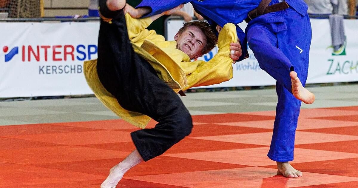 Ein Kampfsportkampf, bei dem ein Mann in einem gelben Gi einen Tritt ausführt, während eine andere Person in Blau ihn festhält.