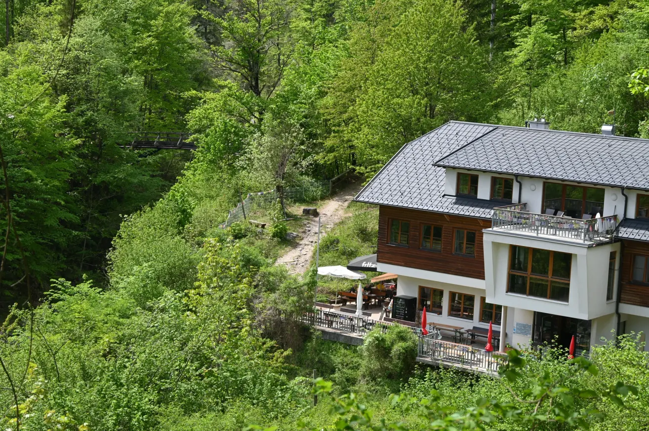A hillside house with a gray roof and brown wooden walls. The house has a balcony with railings and windows. It is surrounded by lush greenery, with a path leading to a bridge.