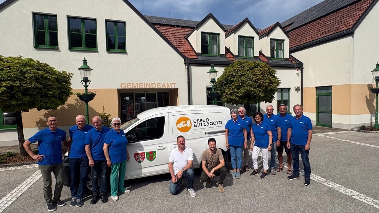 A group of people wearing blue shirts and pants are posing in front of a white van with a logo and the text 'Essen auf radern'. They are standing in front of a building with the sign 'Gemeindeamt' above the entrance.