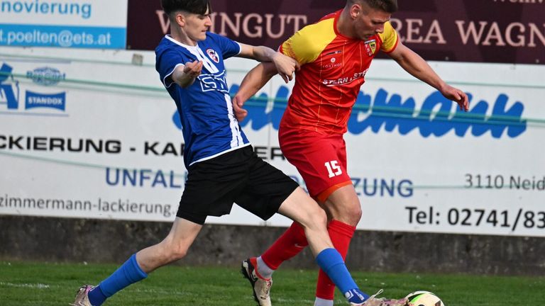 Zwei Fußballspieler konkurrieren auf einem Feld, einer in Blau und der andere in Rot. Der Spieler in Blau versucht, den Spieler in Rot zu blockieren, der das Trikot mit der Nummer 15 trägt.