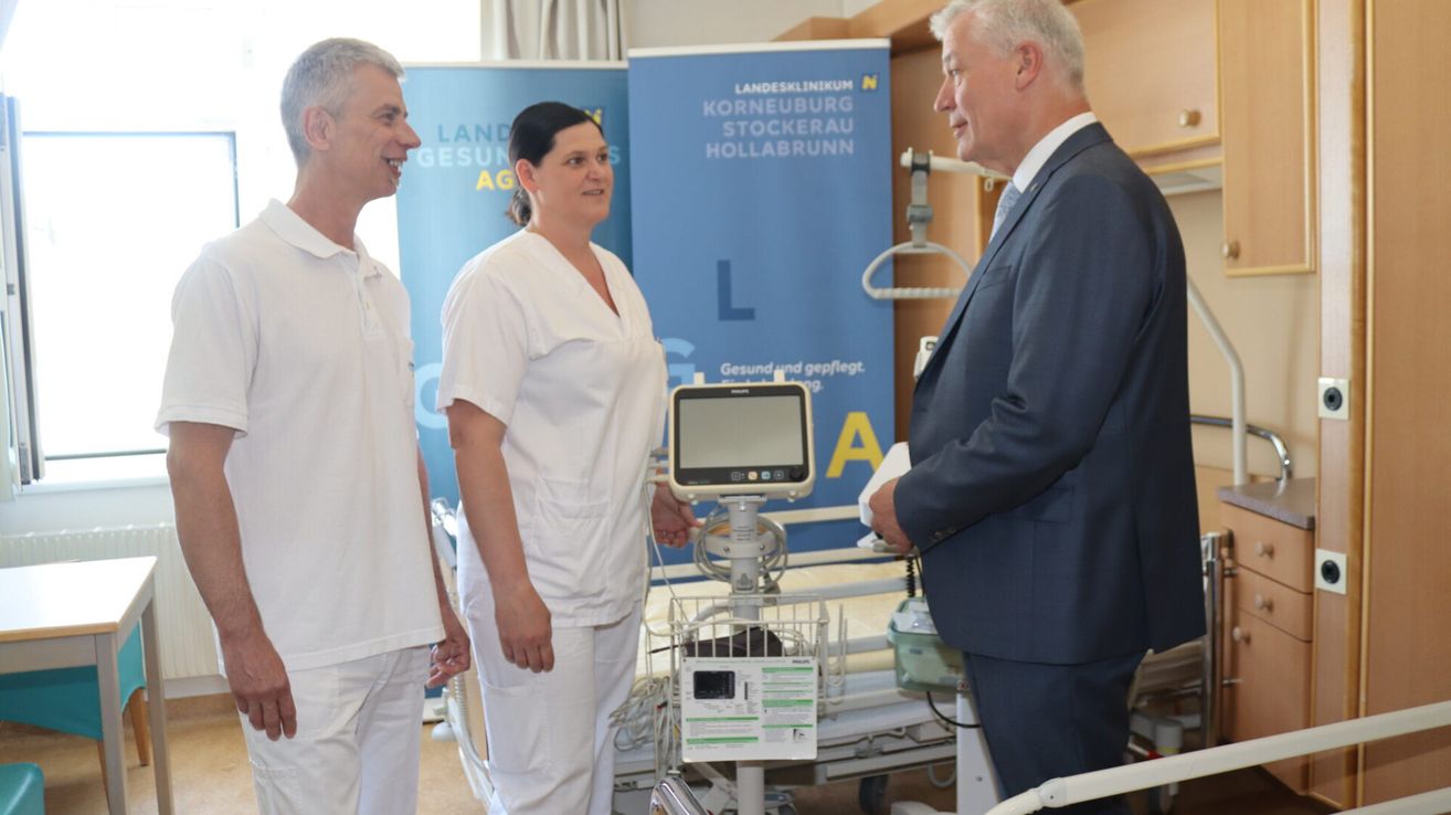 Drei Personen in einem Krankenhauszimmer, eine ist Krankenschwester, die anderen beiden diskutieren etwas in der Nähe eines Krankenhausbettes. Ein blaues Schild steht 'Landesklinikum Korneuburg Stockerau Hollabrunn'.