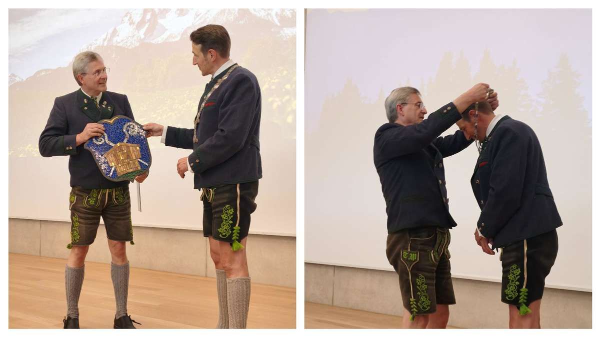 Zwei Männer in traditioneller bayerischer Kleidung stehen auf einer Bühne. Ein Mann hält einen Schild, während der andere dem ersten Mann eine Medaille anlegt.