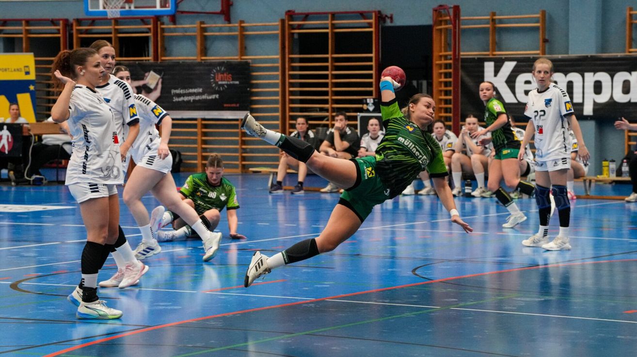 Eine Handballspielerin in einem grünen Trikot springt in der Luft und ist bereit, einen roten Ball zu werfen. Hinter ihr sitzen andere Spieler auf dem Boden und beobachten die Aktion.