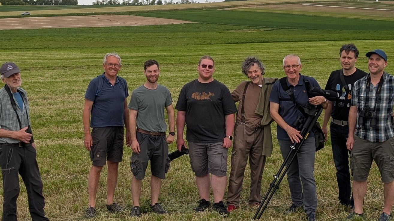 Fünf Männer stehen lächelnd in einem Grasfeld. Sie tragen Freizeitkleidung und einer hat eine Kamera auf einem Stativ.