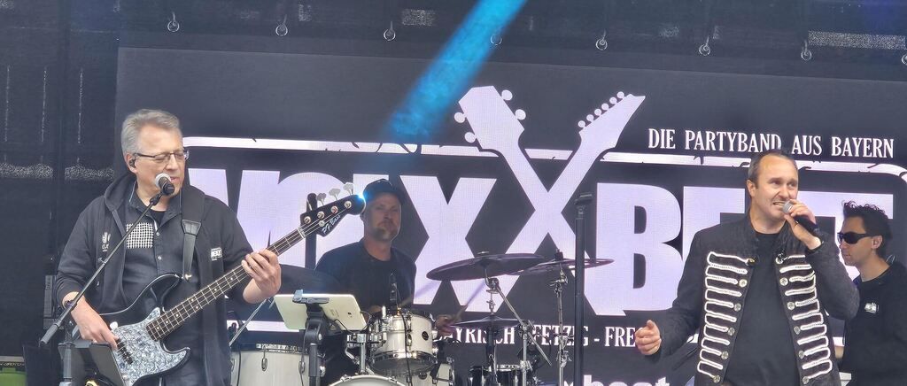Drei Musiker spielen auf der Bühne mit einem Banner, das 'Die Partyband' liest. Einer spielt Gitarre, einer Schlagzeug und einer Akkordeon. Der Schlagzeuger hat ein Mikrofon auf seinem Schlagzeug.