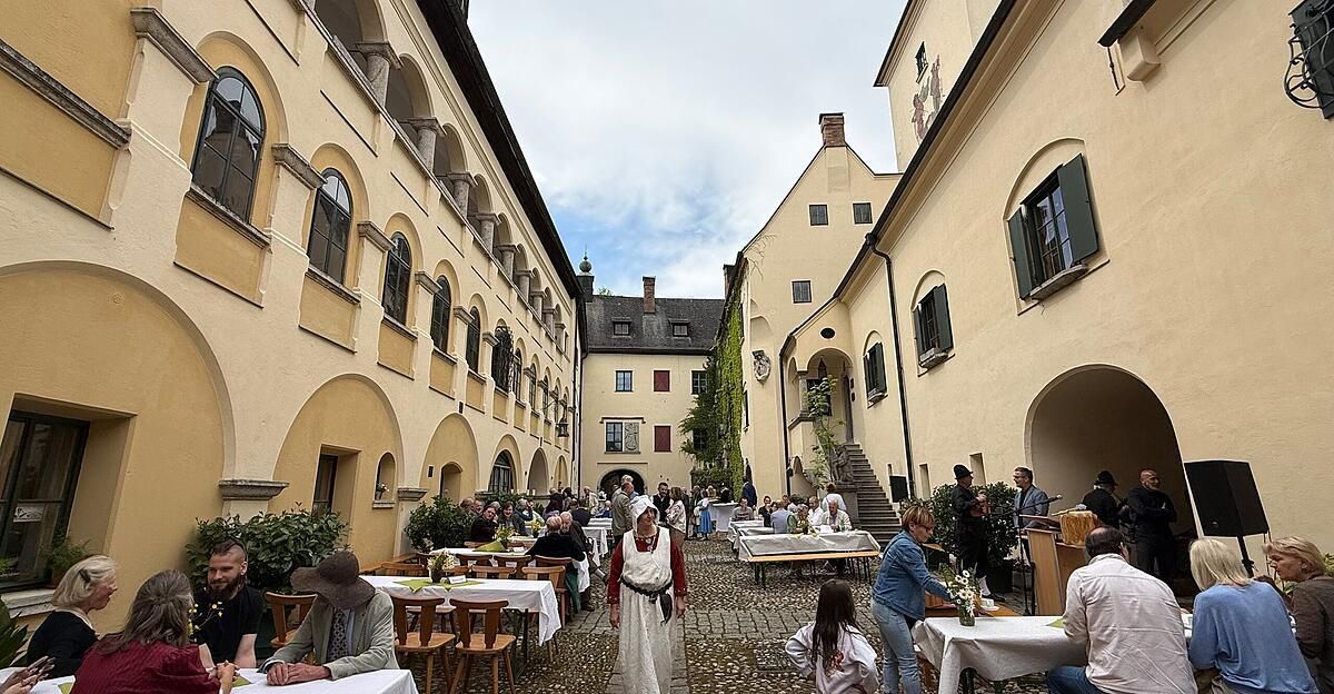 Ein mittelalterlicher Innenhof gefüllt mit Menschen, die an Tischen sitzen, umgeben von historischen Gebäuden. Eine Frau in traditioneller Kleidung geht durch den Hof.