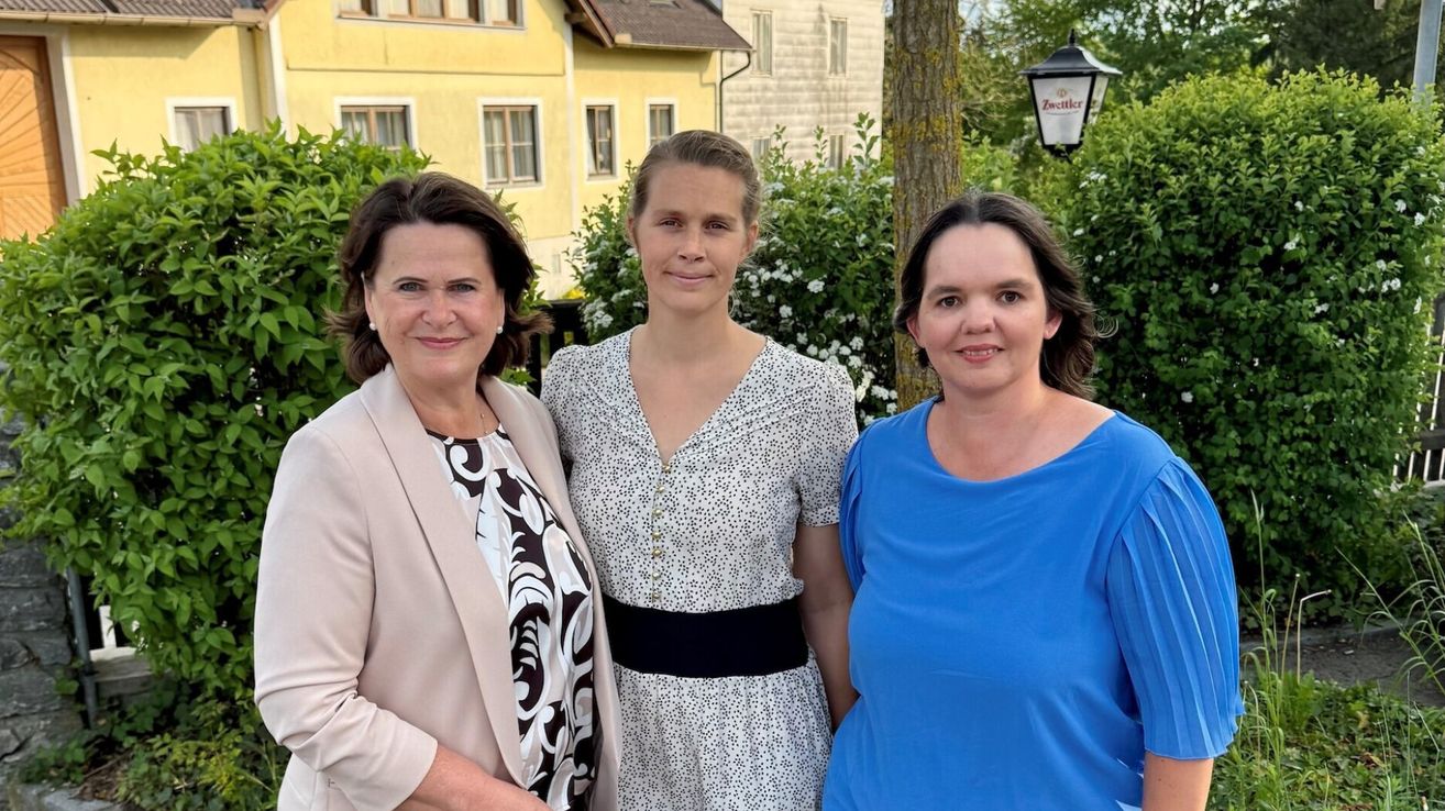Three women stand in front of a yellow building, smiling for the camera. One wears a blue shirt, another a white dress, and the third a beige blazer.