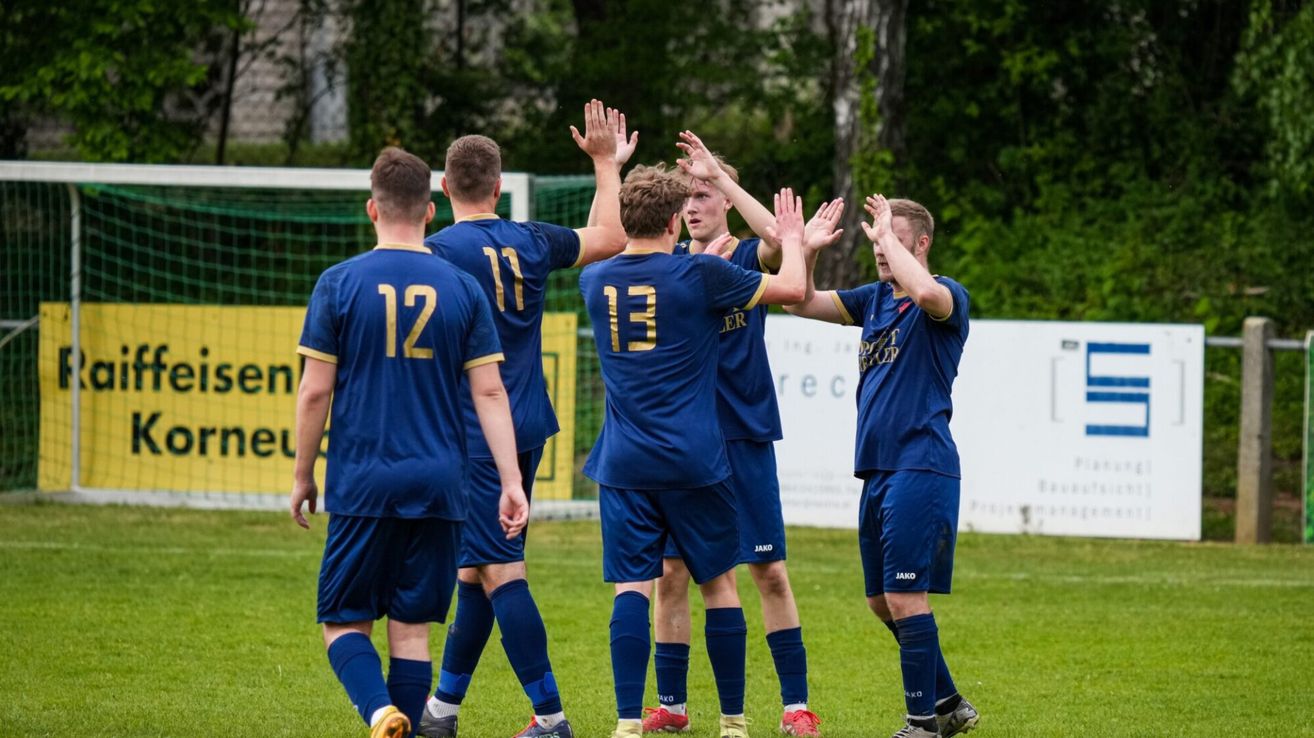 Fünf Fußballspieler in blauen Trikots feiern auf einem Feld, mit den Nummern 12, 17 und 13 auf dem Rücken. Der Spieler mit der Nummer 13 hebt beide Hände. Bäume und ein Zaun sind im Hintergrund.