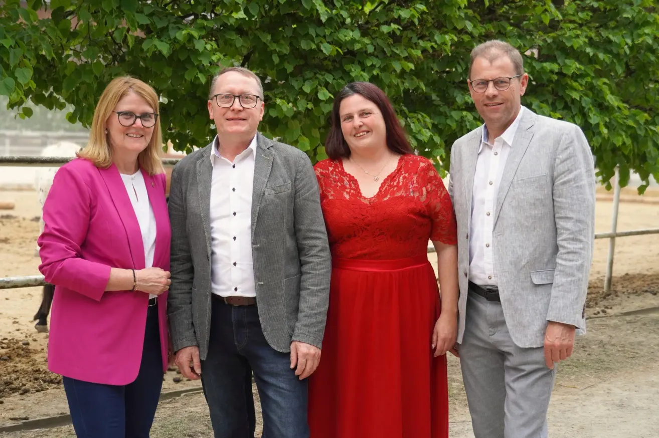Vier Menschen stehen eng beieinander vor einem grünen Baum und lächeln für ein Foto. Eine Frau in einem roten Kleid, ein Mann in einem grauen Jackett und zwei weitere in hellrosa und grauen Anzügen.