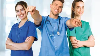 Three smiling medical professionals, a man and two women, point towards the camera. The man wears a stethoscope, and all are in blue and green scrubs.