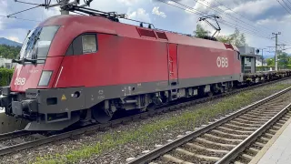 A red train is parked on a track with gravel and grass nearby. The train has a white logo on its side.