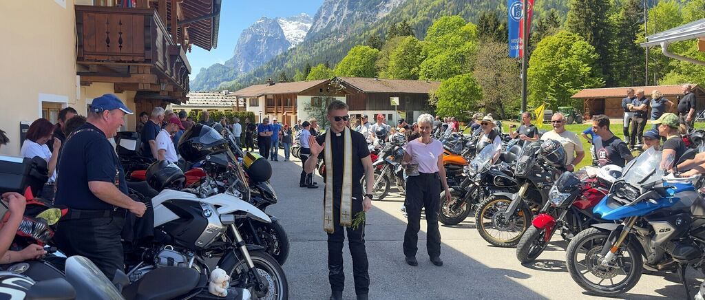 Ein Priester winkt einer Gruppe von Motorradfahrern mit ihren Motorrädern in einer Freiluftfläche zu, mit Gebäuden und Bergen im Hintergrund.