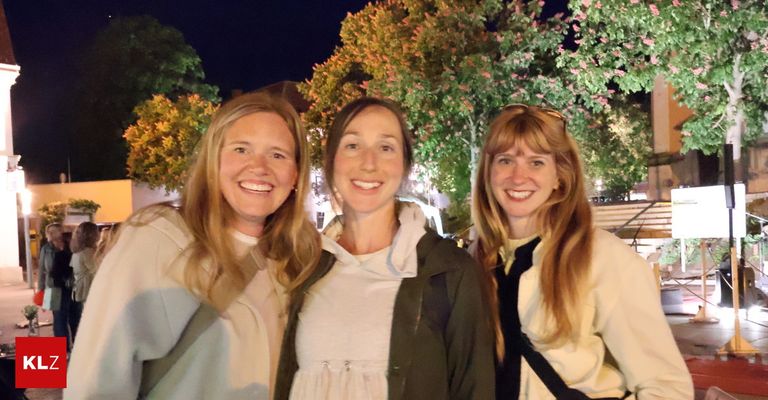 Three women smile for a photo at night under trees with pink flowers and city lights.