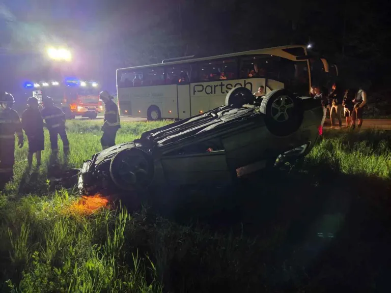 Ein umgekipptes Auto liegt bei Nacht im Gras, ein Bus ist in der Nähe geparkt und Rettungsfahrzeuge sind am Unfallort.