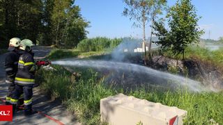 A firefighter sprays water from a hose into a field, aiming to extinguish a fire. Trees and a small building are in the background.
