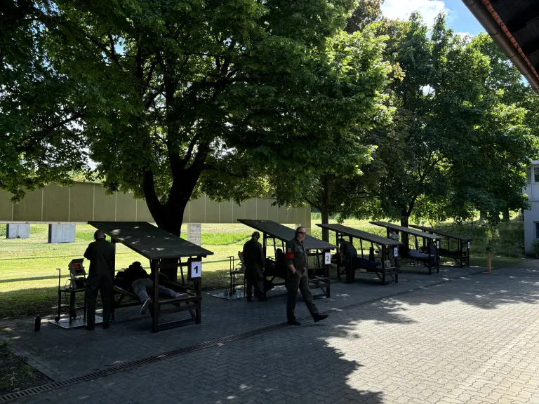 Eine Reihe nummerierter Schießstände mit Personen, die in einem Außenbereich unter Bäumen und einem bewölkten Himmel stehen und gehen.