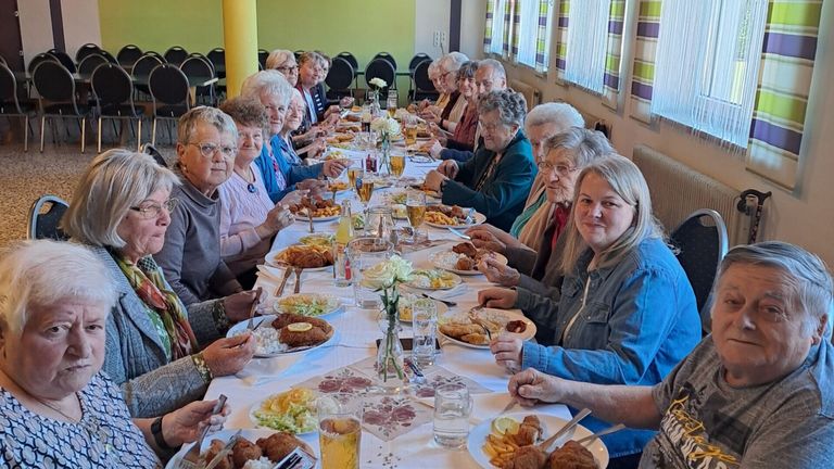 Eine Gruppe älterer Menschen genießt gemeinsam ein Essen an einem langen Tisch, mit verschiedenen Gerichten und Getränken vor ihnen.