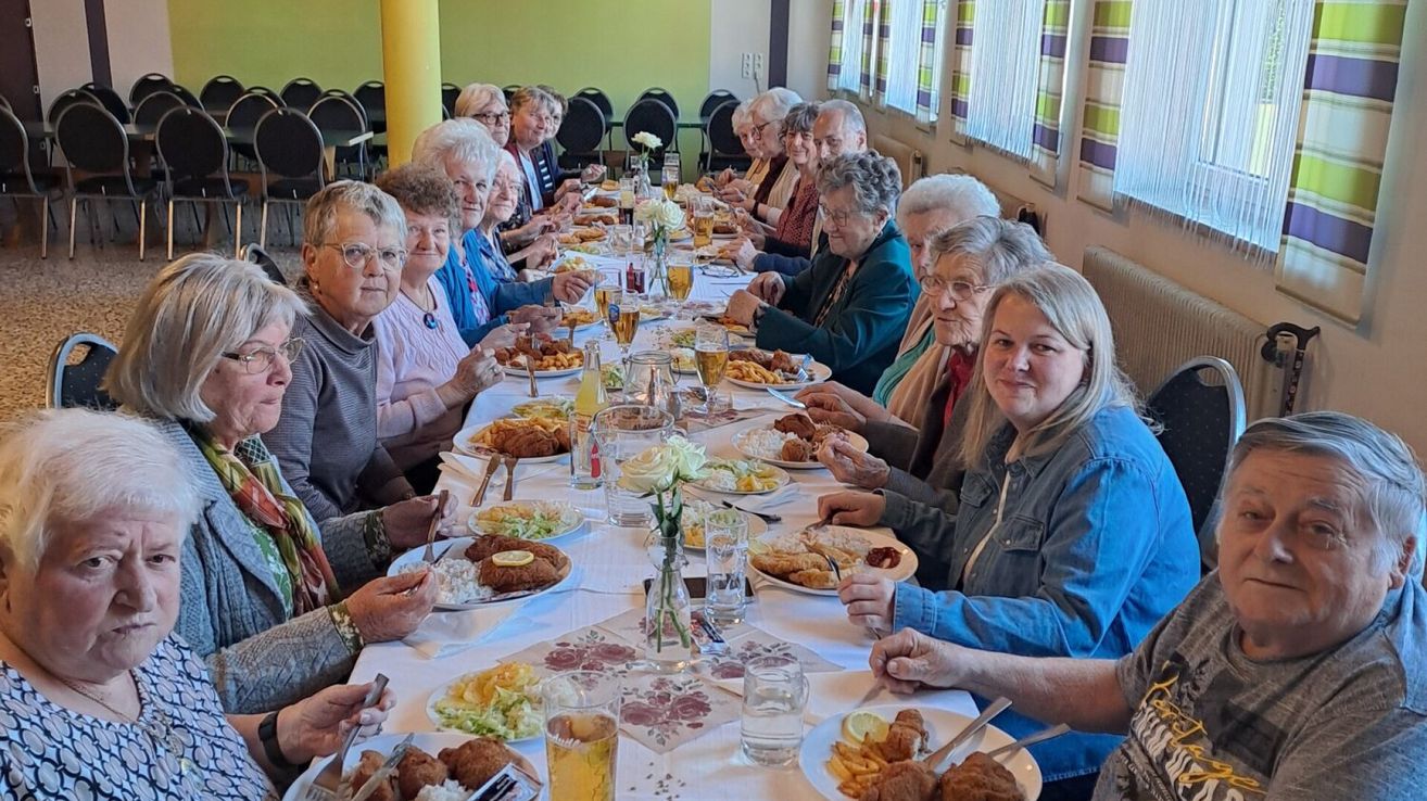 Eine Gruppe älterer Menschen genießt gemeinsam ein Essen an einem langen Tisch, mit verschiedenen Gerichten und Getränken vor ihnen.