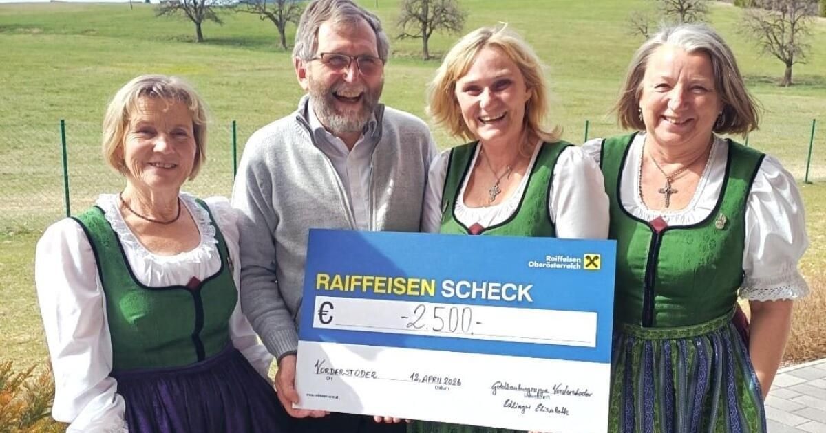 Vier Menschen in traditioneller bayerischer Kleidung halten einen blauen Raiffeisen-Bankscheck über 2.500 Euro. Sie lächeln und posieren für ein Foto auf einer Wiese mit Bäumen im Hintergrund.