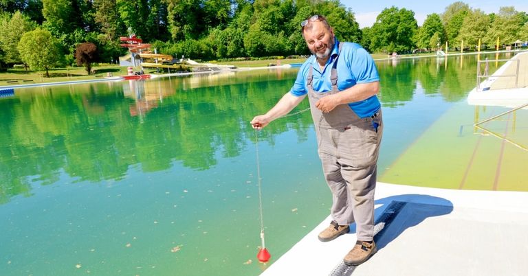 A man in overalls stands by a pool, holding a red float in the water, smiling. In the background, there is a slide, greenery, and a clear blue sky.