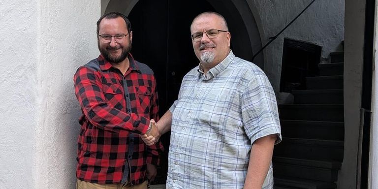 Two men stand next to each other, smiling, with one holding a bracelet, both wearing glasses and standing in front of a building.