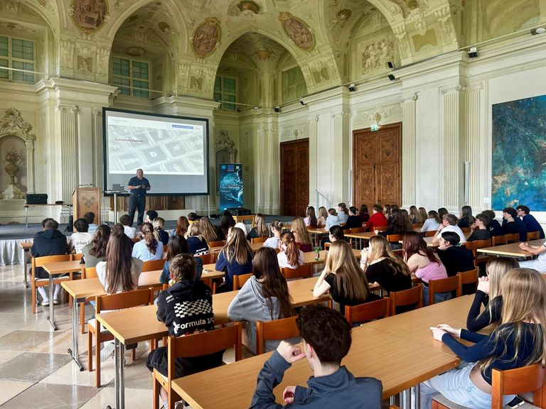 Ein Mann präsentiert eine Folie auf einer Leinwand in einem Hörsaal vor einer Gruppe von Studenten, die an Tischen sitzen.