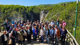A group of people pose on a wooden bridge with a waterfall in the background. They are all smiling and some are waving. The waterfall is surrounded by greenery.