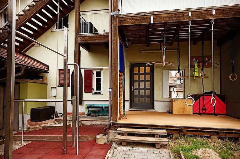 Ein Outdoor-Fitnessstudio mit Kletterseilen und Ringen auf einer Holzveranda mit Fliesenboden. Ein Haus mit roten Fensterläden und einem Balkon mit Geländer.