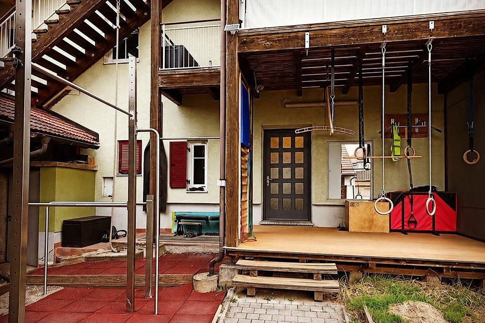 Ein Outdoor-Fitnessstudio mit Kletterseilen und Ringen auf einer Holzveranda mit Fliesenboden. Ein Haus mit roten Fensterläden und einem Balkon mit Geländer.