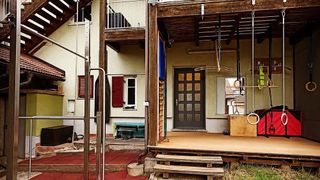 An outdoor gym with climbing ropes and rings on a wooden porch with a tiled floor. A house with red shutters and a balcony with a railing.