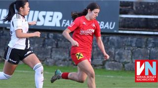 Two female soccer players in red and black uniforms are running on a field. A stone wall is behind them with a banner that reads USER FA.