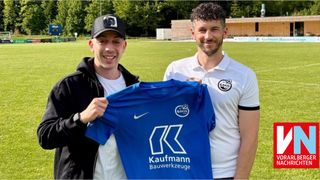 Two men pose on a soccer field, one holding a blue Kaufmman Bauwerkzeuge jersey. The man on the left wears a black cap and jacket. Behind them is a grassy field with a fence and trees.