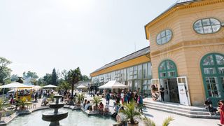 A large crowd gathers around a fountain outside a yellow building with arched windows, on a sunny day.