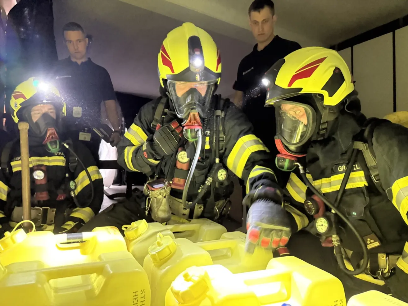 Two firefighters in full gear are seated, each handling equipment. A third firefighter stands behind them, also equipped. They are surrounded by yellow containers.
