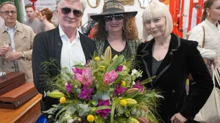 Three people stand close to each other, smiling, holding a large bouquet of flowers. They wear sunglasses and jackets. In the background, people and decorations are visible.