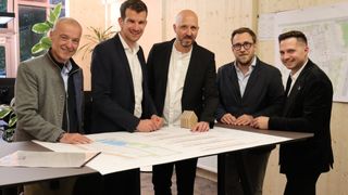 Four men in suits discuss over a large blueprint on a table, one holding a small wooden house model.