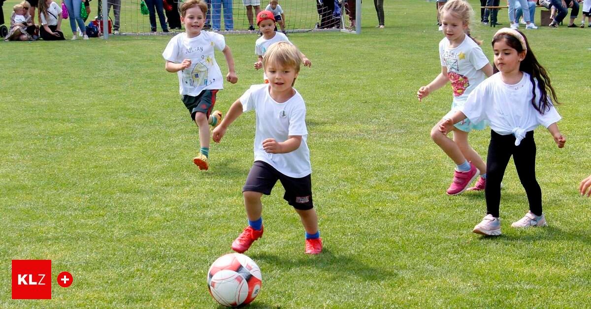 Several children play soccer on a grassy field. A boy in red shoes kicks the ball. Other children run around, some wearing hats. A soccer goal is in the background.