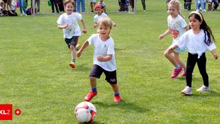 Several children play soccer on a grassy field. A boy in red shoes kicks the ball. Other children run around, some wearing hats. A soccer goal is in the background.