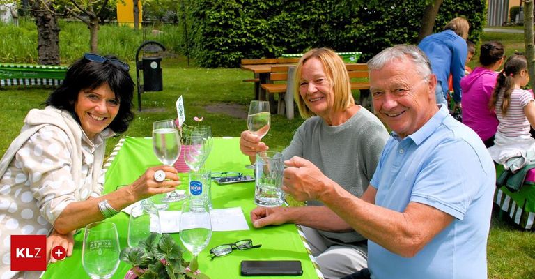 Three people sit around a table, smiling and holding wine glasses. The table is covered with a green tablecloth and has flowers, a phone, and eyeglasses on it. There are benches and trees in the background.