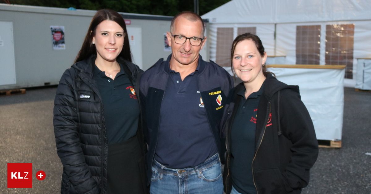 Drei Menschen stehen draußen in der Nähe eines Zeltes. Der Mann trägt eine Feuerwehrjacke und eine Brille. Die Frauen tragen Jacken und lächeln.