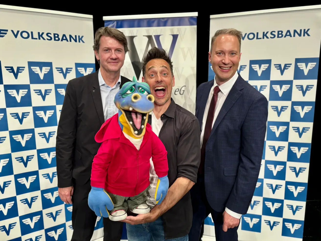Drei Männer und eine Puppe posieren für ein Foto, alle lächeln. Der Mann auf der linken Seite trägt einen schwarzen Anzug. Der Mann in der Mitte hält eine blaue Puppe mit einem roten Jackett. Der Mann auf der rechten Seite trägt einen dunkelblauen Anzug mit Krawatte. Hinter ihnen sind Banner mit Logos.