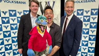 Three men and a puppet posing for a picture, all smiling. The man on the left wears a black suit. The man in the middle holds a blue puppet with a red jacket. The man on the right wears a dark blue suit with a tie. Behind them are banners with logos.