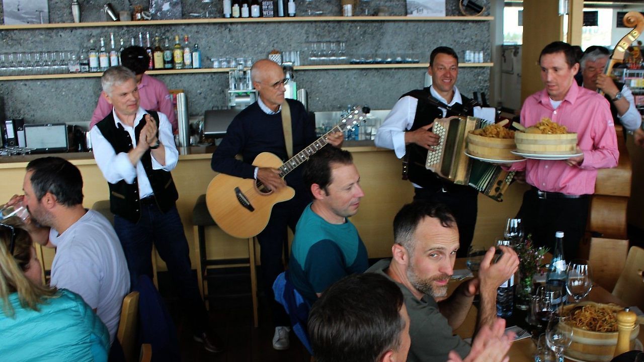 A band plays in a bar with patrons sitting. A man plays guitar, another plays accordion. A man in a blue shirt sits in the front.