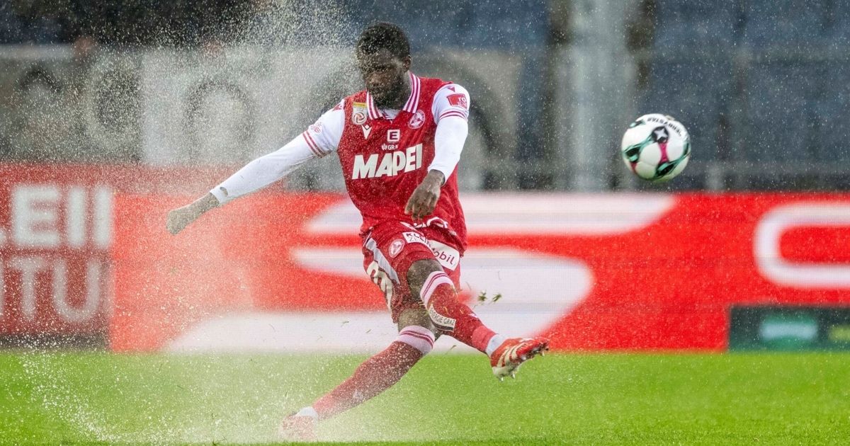 A soccer player in a red uniform with 'Mapei' on it kicks a ball during a match. The field is wet with splashes around the player.