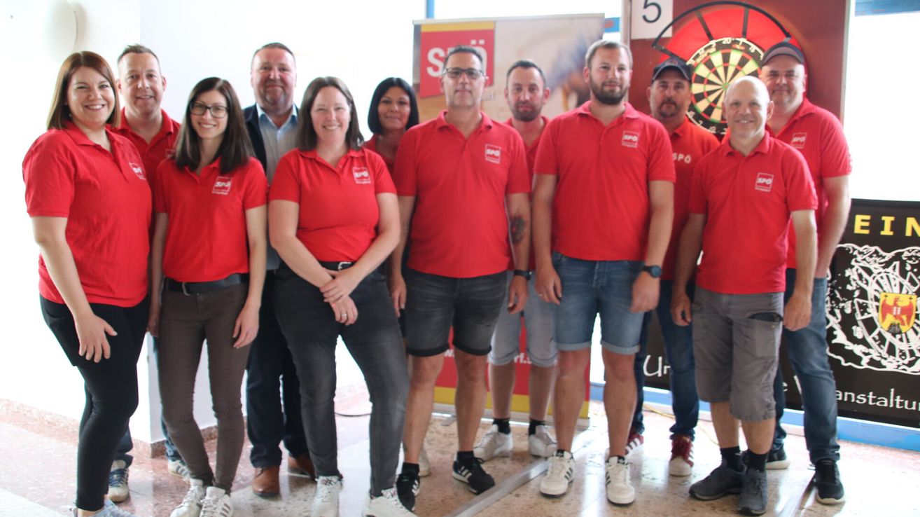 A group of adults in matching red shirts and various colored pants are posing for a photo in front of a dartboard and banner.