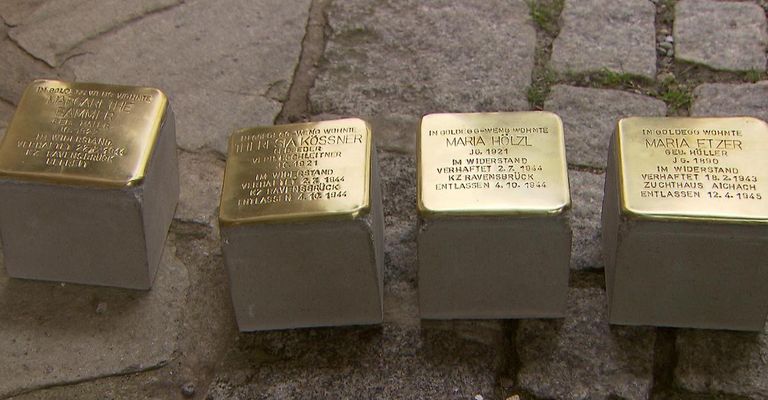 Four gold plaques on concrete blocks, each with names and dates, lie on a ground. Two plaques read 'Maria Holzl' and 'Im Widerstand verhaftet'. Another plaque reads 'Gia Kosssner' and 'Im Widerstand verhaftet'. The fourth plaque is partially visible.