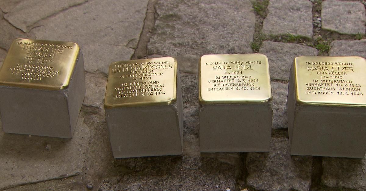 Four gold plaques on concrete blocks, each with names and dates, lie on a ground. Two plaques read 'Maria Holzl' and 'Im Widerstand verhaftet'. Another plaque reads 'Gia Kosssner' and 'Im Widerstand verhaftet'. The fourth plaque is partially visible.